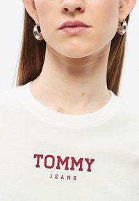 Λευκό φούτερ με στρογγυλή λαιμόκοψη και λογότυπο "TOMMY JEANS" σε βουργουνδί χρώμα. Το μοντέλο φορά μεγάλα ασημένια κρίκους σκουλαρίκια σε ουδέτερο φόντο.