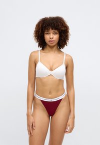 Hvit bh med tynne stropper og formede kopper, kombinert med burgunder stringtruse med en hvit kant med "TOMMY HILFIGER"-logo. Glatt stoffstruktur.