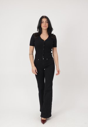 Femme aux longs cheveux foncés portant un haut noir boutonné, un pantalon évasé noir et des talons pointus bordeaux, debout devant un fond blanc.