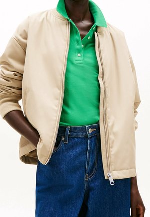 Person wearing a beige zip-up jacket over a green collared shirt and blue jeans, with one hand in jacket pocket.