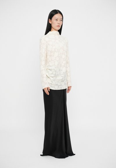 Top a maniche lunghe e collo alto in crema chiaro con texture floreale, abbinato a una gonna nera lunga fino al pavimento con un design elegante e fluente.