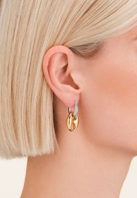 Los pendientes de aro en oro y plata presentan un óvalo de oro pulido con un diseño recortado, acentuado por una fila de pequeñas piedras transparentes.