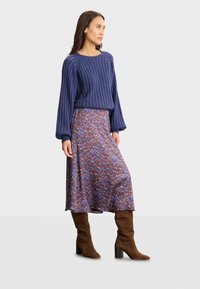 Marineblaue gerippte Strickjacke mit Puffärmlen, kombiniert mit einem Midirock mit einem mehrfarbigen geometrischen Muster und kniehohen braunen Wildlederstiefeln.