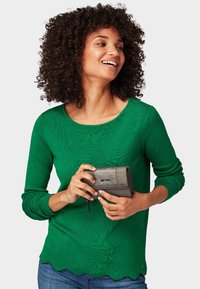 Maglione a coste verde con orlo scalloppato, abbinato a un portafoglio grigio. Il modello tiene il portafoglio ed indossa jeans.
