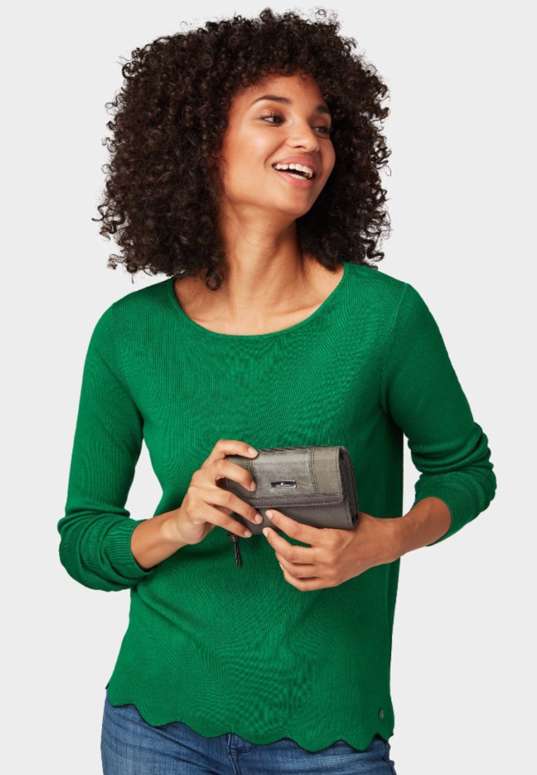 Grüner gerippter Strickpullover mit gewelltem Saum, kombiniert mit einer grauen Geldbörse. Das Model hält die Geldbörse und trägt Jeans.