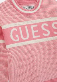 Rózsaszín kötött pulóver csíkos mintával, fehér vízszintes sávval, amelyen a "GUESS" logó vastag betűkkel szerepel.