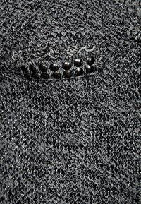 Černá pletená látka s texturovaným vzorem a dekorativními černými korálky podél tkaného pásku. Má mírný lesk.