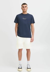 Navy blue cotton t-shirt with "Bonjour Côte d'Azur" text, paired with light-colored, textured shorts and black-and-white sneakers.