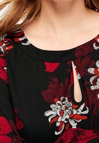 Gros plan sur un chemisier noir avec des motifs floraux rouges, blancs et bordeaux, et un petit détail d'encolure en forme de trou de serrure.