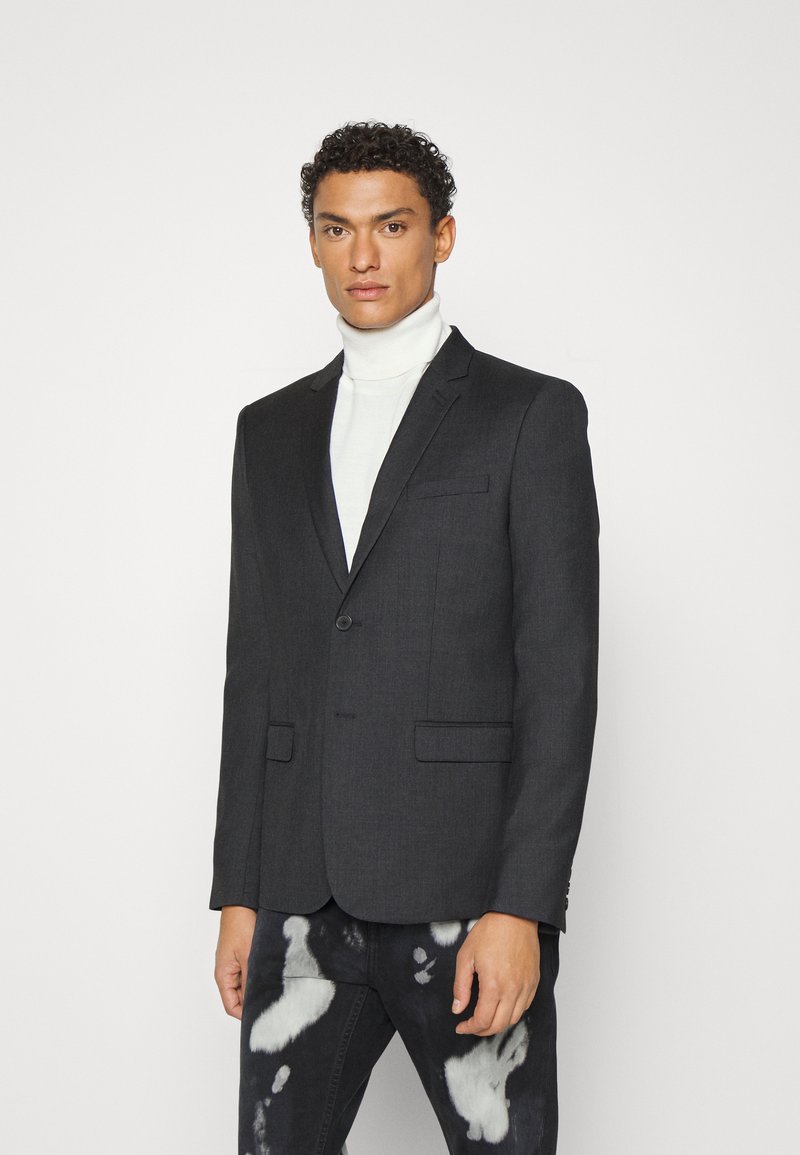 Dunkelgrauer Blazer mit taillierter Passform, einem Einzelknopfverschluss und zwei Fronttaschen, getragen über einem weißen Rollkragenpullover.