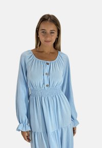 Robe bleu clair à manches longues avec une taille smockée, un col rond et trois boutons sur le devant. Tissu doux et texturé avec un design en couches.