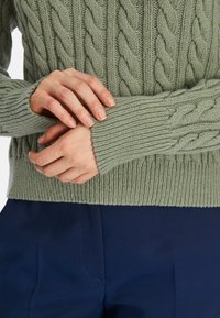 Strickpullover in sanftem, gedämpften Grün mit strukturiertem Kabeldesign und gerippten Bündchen. Kombiniert mit navyblauen Hosen.