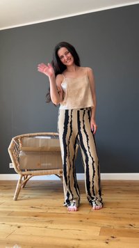 Crop top beige lucido abbinato a pantaloni a vita alta a righe nere e crema. La modella si trova su un pavimento in legno, mentre saluta. Sedia in rattan sullo sfondo.