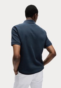 Camisa polo de textura acanalada en azul marino oscuro, con un cuello corto y mangas enrolladas, combinada con pantalones de color claro.