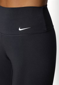 Leggings noirs taille haute en tissu extensible, arborant un logo Nike blanc sur le devant, avec des coutures lisses et une coupe ajustée.