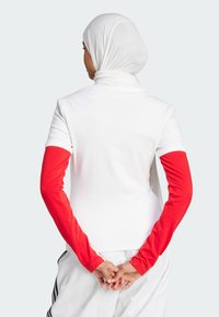 Bela rebrasta majica s kratkimi rokavi in dolgimi rdečimi rokavi, značilna z ohlapnim krojem. Oseba nosi bel hijab.