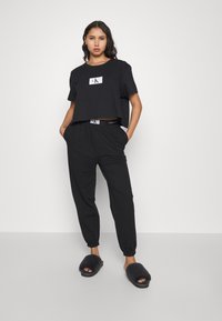 T-shirt noir court avec un logo blanc, assorti à un pantalon noir fuselé avec une taille élastique et des poches, complété par des chaussons glissants en fourrure.