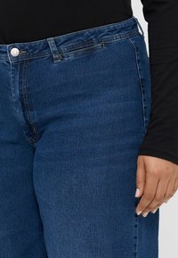 Jeans de denim azul-escuro com um design de perna reta. Apresentam um fecho de botão e detalhes de costura discretos ao longo dos bolsos e da cintura.