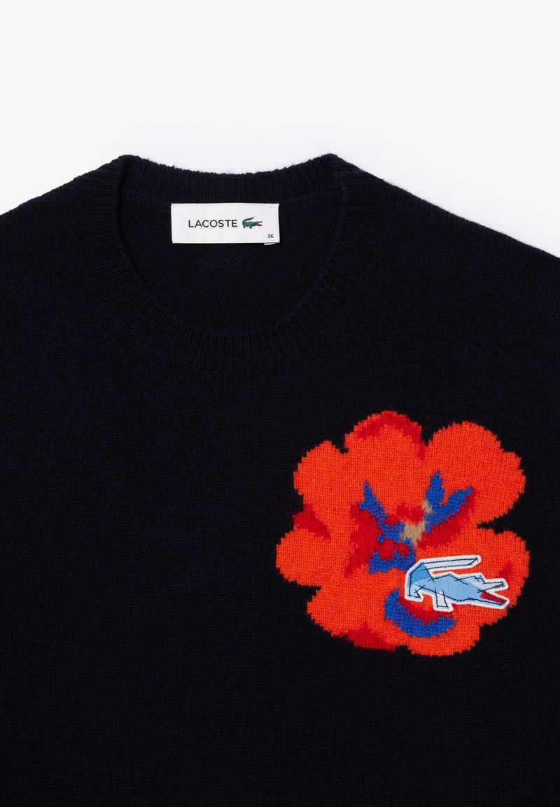 Marineblauer Strickpullover mit einem großen roten Blumenmotiv und blauen Akzenten sowie einem kleinen gestickten Logo in der Nähe der Blume.