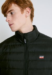 Chaqueta negra tipo puff con textura acolchada vertical, cuello alto y logo de Levi's en el lado izquierdo del pecho. Cierre de cremallera para mayor funcionalidad.