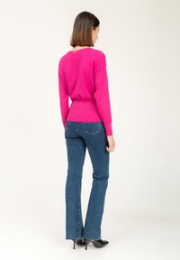 Fuchsia trui met geribde zoom en lange mouwen, gecombineerd met blauwe uitlopende jeans. Zwarte puntige schoenen en een eenvoudig halslijn detail zijn zichtbaar.