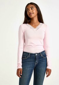 Långärmad rosa topp med V-ringning och spetskant. Matchas med blå denimjeans med vita sömdetaljer.