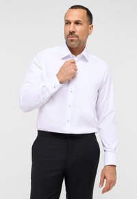 Hvid skjorte med en subtil tekstureret mønster, der har en spredekrave, knaplukning foran og mansjetter. Matchet med sorte bukser.