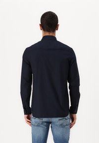 Cămașă albastru închis cu mâneci lungi, cu manșete cu nasturi și yoke la spate. Purtați cu blugi deschiși la culoare, văzuți din spate pe un fundal alb.