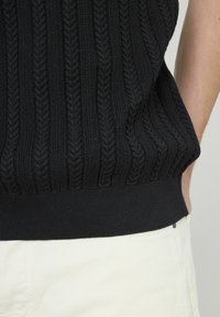 Top sans manches en tricot noir présentant un motif de câble texturé et un ourlet côtelé uni. Associé à un pantalon de couleur claire.