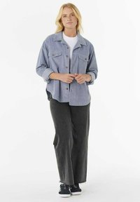 Giacca camicia di corduroy azzurro chiaro con due tasconi sul petto, bottoni e maniche arrotolate, abbinata a pantaloni grigi a gamba larga e sneakers nere.