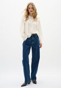 Blusa bianca a maniche lunghe con bordi arricciati e ricami, abbinata a jeans in denim blu a vita alta. Scarpe nere a punta completano il look.