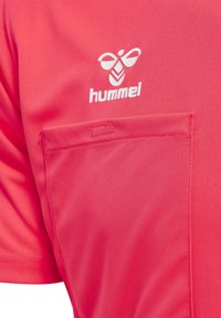 Camiseta deportiva rosa brillante con mangas cortas, bolsillo delantero y logo blanco de "hummel" con un gráfico estilizado encima.