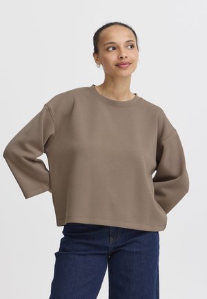 Felpa marrone oversize con maniche ampie e scollo rotondo, realizzata in materiale morbido, abbinata a jeans di denim blu scuro.