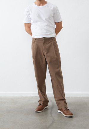 Mann trägt weißes kurzärmeliges T-Shirt, braune Hose und braune Schuhe, steht mit den Händen hinter dem Rücken auf grauem Boden und weißer Wand im Hintergrund.