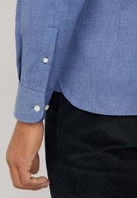 Chemise boutonée texturée bleu clair avec un ourlet courbé, dotée de boutons blancs aux poignets et d'une coupe ajustée. Pantalon en velours côtelé noir.
