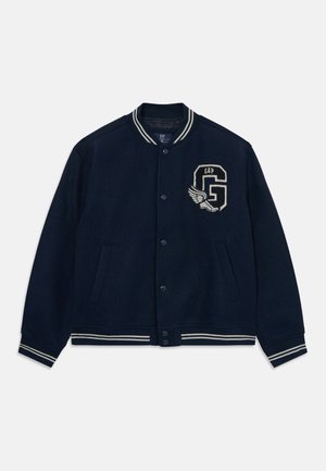 Chaqueta varsity de color azul marino con puños y cuello a rayas, botones a presión, bolsillos laterales y un parche "GAP" con el logo de una zapatilla alada en el pecho.