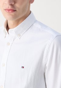 Camicia bianca con colletto a punta, tessuto strutturato e un piccolo logo ricamato in rosso, bianco e blu sul petto.