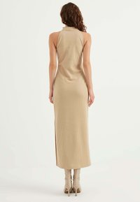 Abito lungo in maglia a coste beige con colletto alto e spacco laterale, caratterizzato da un design aderente e hardware minimale. Abbinato a stivali al ginocchio.