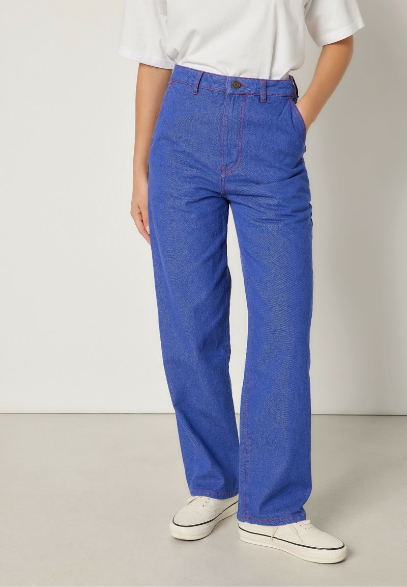 Blå denimbyxor med vida ben, synlig röd sömnad, hög midja och sidfickor, parat med vita sneakers.