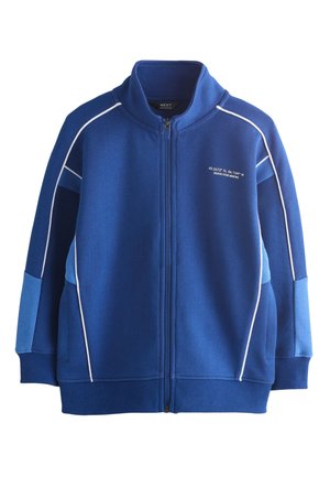 Barn med krøllet hår iført en blå lynlåstrøje og matchende shorts med hvide sneakers og stribede sokker.