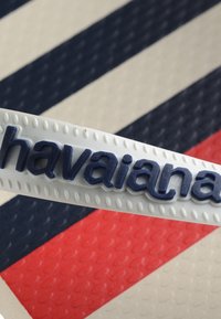 Bracelet en caoutchouc blanc avec texte en relief bleu foncé « havaianas », sur un fond texturé avec des rayures diagonales bleu marine, rouge et crème.