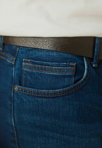 Vaqueros de mezclilla en azul oscuro con costuras naranjas y un pequeño bolsillo trasero, combinados con un cinturón de piel negra texturizada.