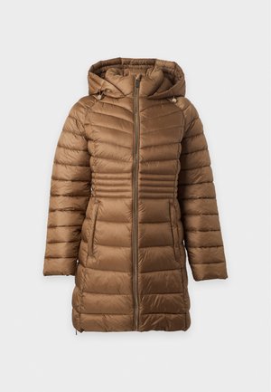 Femme portant un manteau matelassé marron à capuche par-dessus une chemise rose boutonnée, un pantalon large gris clair et des chaussures blanches à talons, debout, droite.