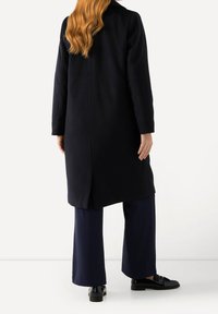 Cappotto in ovatta blu navy con taglio dritto, spacco posteriore singolo e maniche lunghe. Abbinato a pantaloni larghi blu navy e mocassini neri.