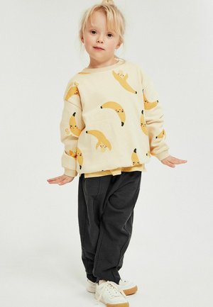 Jong kind staat met licht uitgestrekte armen, draagt een crèmekleurige trui met lachende banaanprints, zwarte broek en witte sneakers.