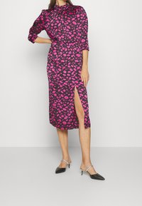 Aansluitende jurk van zwarte stof met een roze lippenprint, lange mouwen, hoge halslijn en een zijsplit. Gecombineerd met zwarte hoge hak mules.