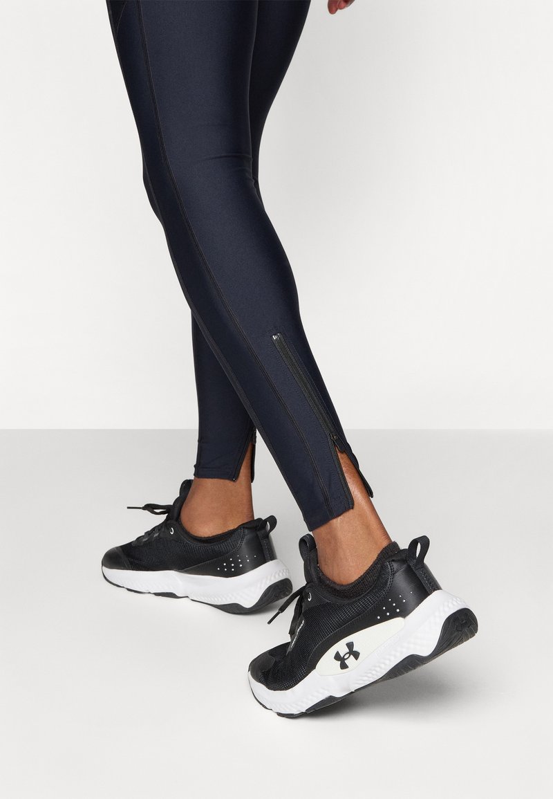 Donkerblauwe legging met zijritsen, gemaakt van een gladde stof, gecombineerd met zwarte sportschoenen met witte accenten en een gestructureerde zool.