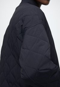 Teget prošivena jakna s dijamantnim uzorkom, s rebrastim ovratnikom i laganom, glatkom teksturom. Prikaz detalja na rukavu u krupnom planu.