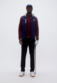 Άνδρας στέκεται κρατώντας μπαστούνι γκολφ, φοράει μπλε γιλέκο, μπορντό πουλόβερ, μαύρο παντελόνι, λευκό γάντι γκολφ, μπλε καπέλο και λευκά αθλητικά παπούτσια.