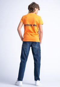 Camiseta de algodón naranja con el texto negro "ETROL IND." en la parte de atrás, combinada con pantalones cargo de denim azules y zapatillas blancas.